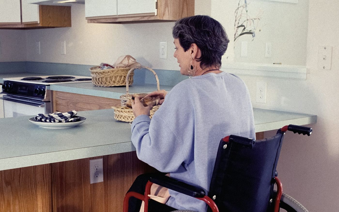 A woman in a wheelchair pulled up to a kitchen counter, with wood-trimmed cabinets and an electric range behind her
