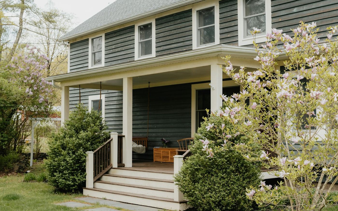 A dark green clapboard house with white-trimmed double-hung windows, a covered front porch and a magnolia in bloom