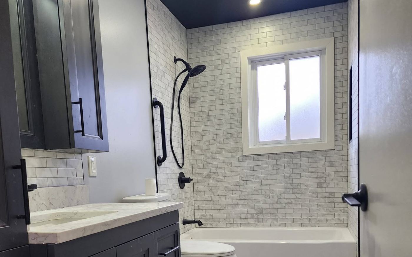 A renovated bathroom with a marble-look subway tiled tub surround, matte black shower fittings, a dark shaker vanity with a stone top, a tall linen cabinet and pale wood-look plank flooring.