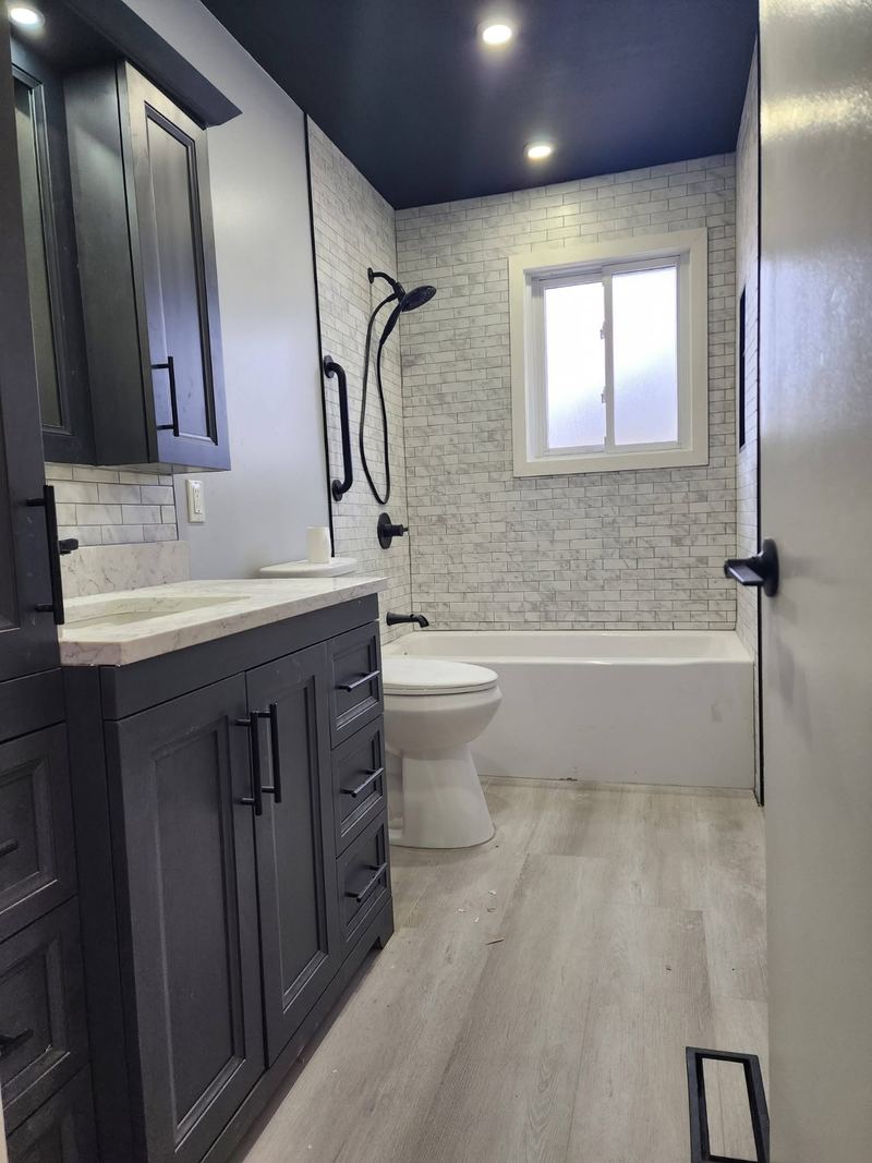A renovated bathroom with a marble-look subway tiled tub surround, matte black shower fittings, a dark shaker vanity with a stone top, a tall linen cabinet and pale wood-look plank flooring.
