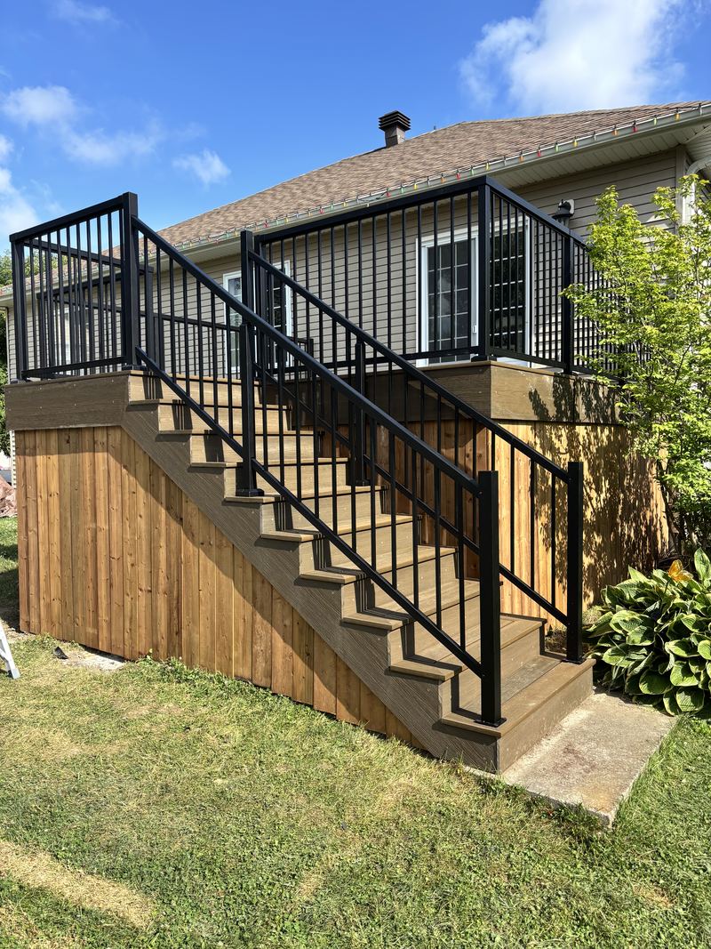 A raised pressure-treated deck against a bungalow, with black aluminum railings, a wide flight of stairs railed on both sides and vertical board skirting below the deck frame.
