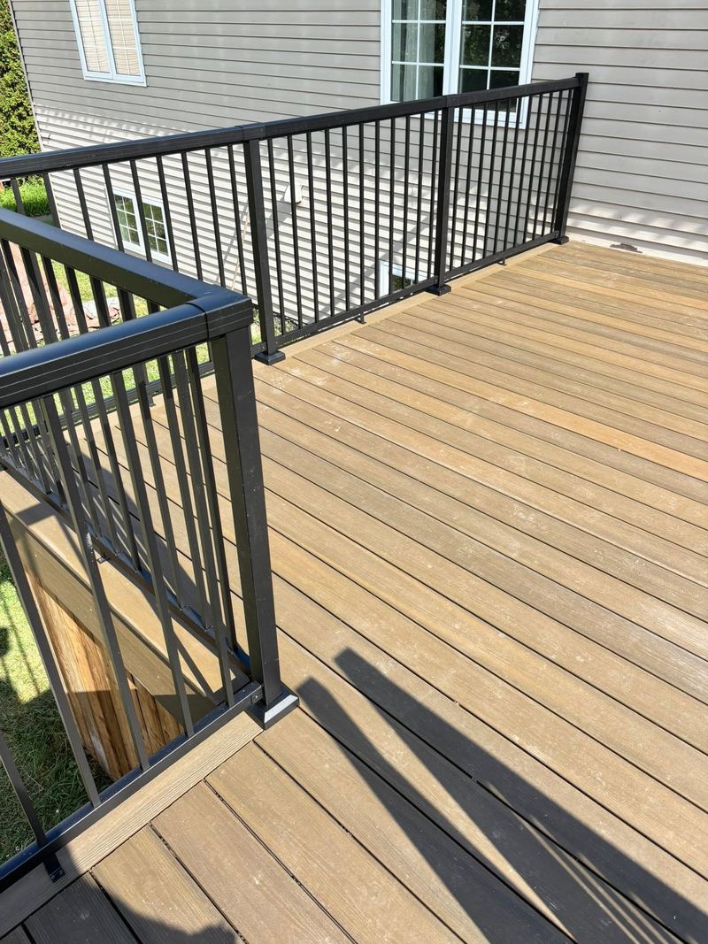 A composite deck surface in warm brown boards with a picture-framed border, black aluminum railings and stairs descending at one corner.