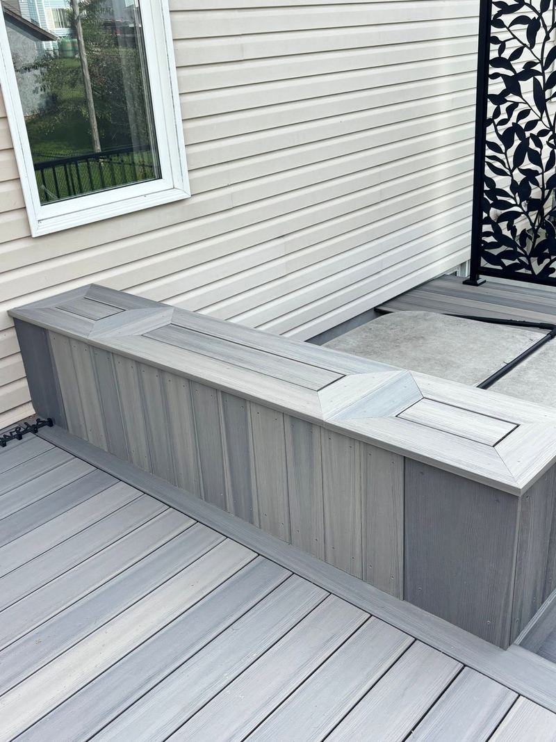 A grey composite deck with a built-in storage bench, mitred picture-frame lids and a black decorative privacy screen against cream siding.