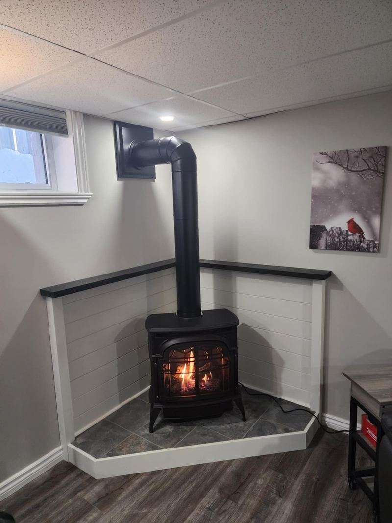 A corner stove alcove finished in white shiplap with a black capped ledge running around it, a tiled hearth pad edged in mitred white trim, and a black stove with the fire lit.