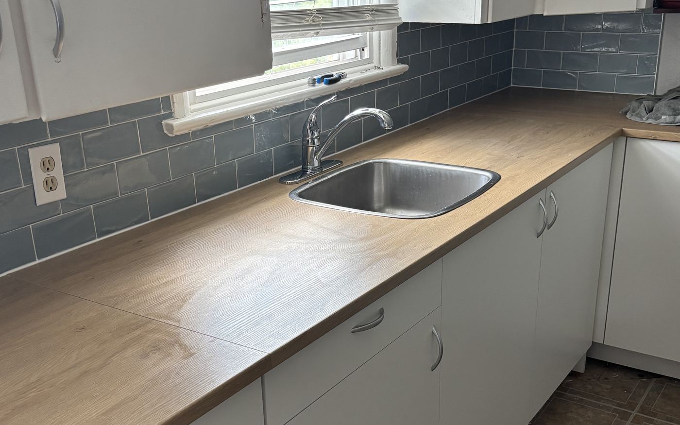 A kitchen mid-renovation with a newly fitted wood-look laminate counter, a stainless steel sink and chrome faucet, and a blue-grey subway tile backsplash above white cabinets.