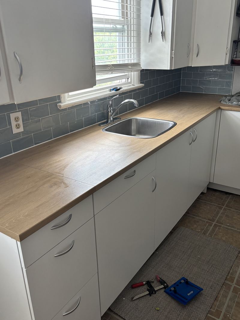 A kitchen mid-renovation with a newly fitted wood-look laminate counter, a stainless steel sink and chrome faucet, and a blue-grey subway tile backsplash above white cabinets.
