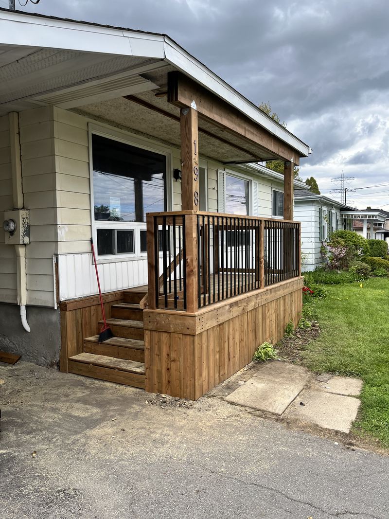 A pressure-treated front porch with a shed-style roof carried on two timber posts, black aluminum balusters between wooden rails, board skirting and open steps down to the driveway.