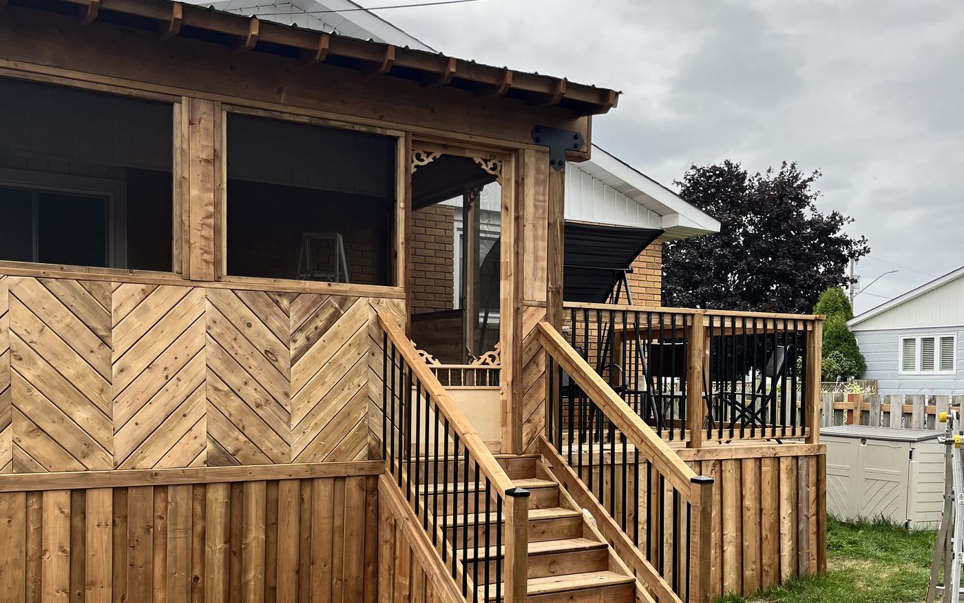 A cedar-toned screened porch with a shingled roof, decorative scrolled corner brackets and skirting laid in a herringbone pattern, with wide stairs and an adjoining open deck with black aluminum balusters.
