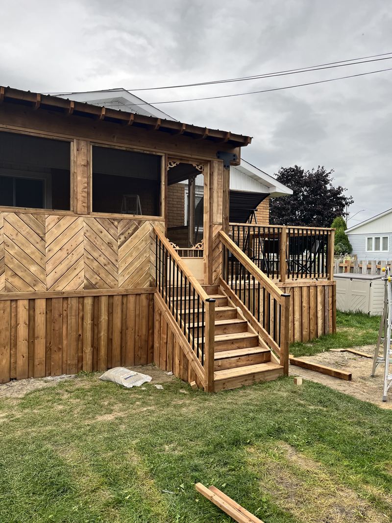 A cedar-toned screened porch with a shingled roof, decorative scrolled corner brackets and skirting laid in a herringbone pattern, with wide stairs and an adjoining open deck with black aluminum balusters.