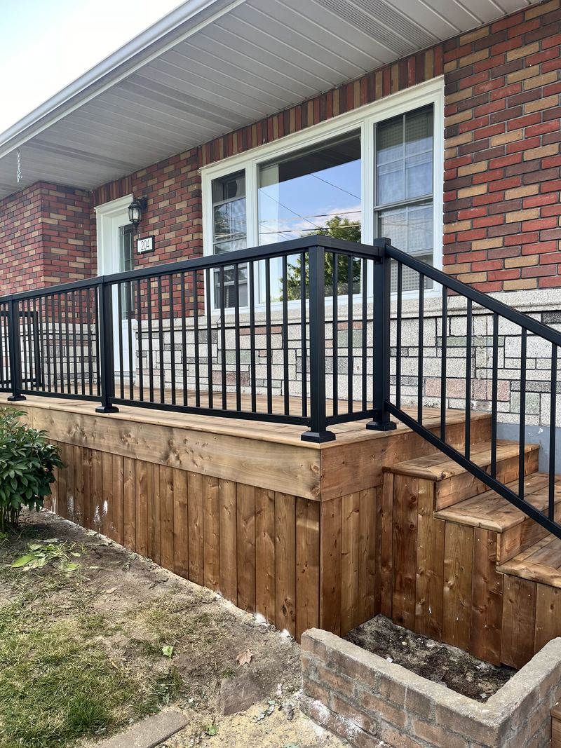 A pressure-treated front veranda on a red brick bungalow, with a black aluminum railing, a wide mitred wooden cap and vertical board skirting, and stairs railed to match.
